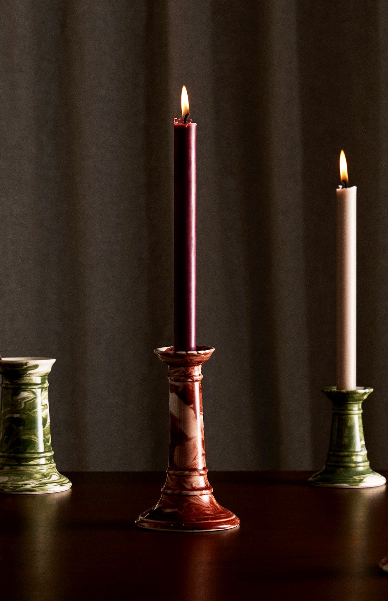 Three Marbled Wardour Candlesticks by THE CONRAN SHOP—one red with a lit burgundy candle, two green (one with an unlit white candle)—stand on a dark surface before a softly lit curtain. Each candlestick features hand-marbled glaze.