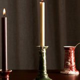 Three tall candles stand on a dark wooden surface. Two are lit—one beige in THE CONRAN SHOP’s Marbled Wardour Candlestick with hand-marbled glaze, and one dark brown in a red holder. A neutral, textured curtain forms the background.