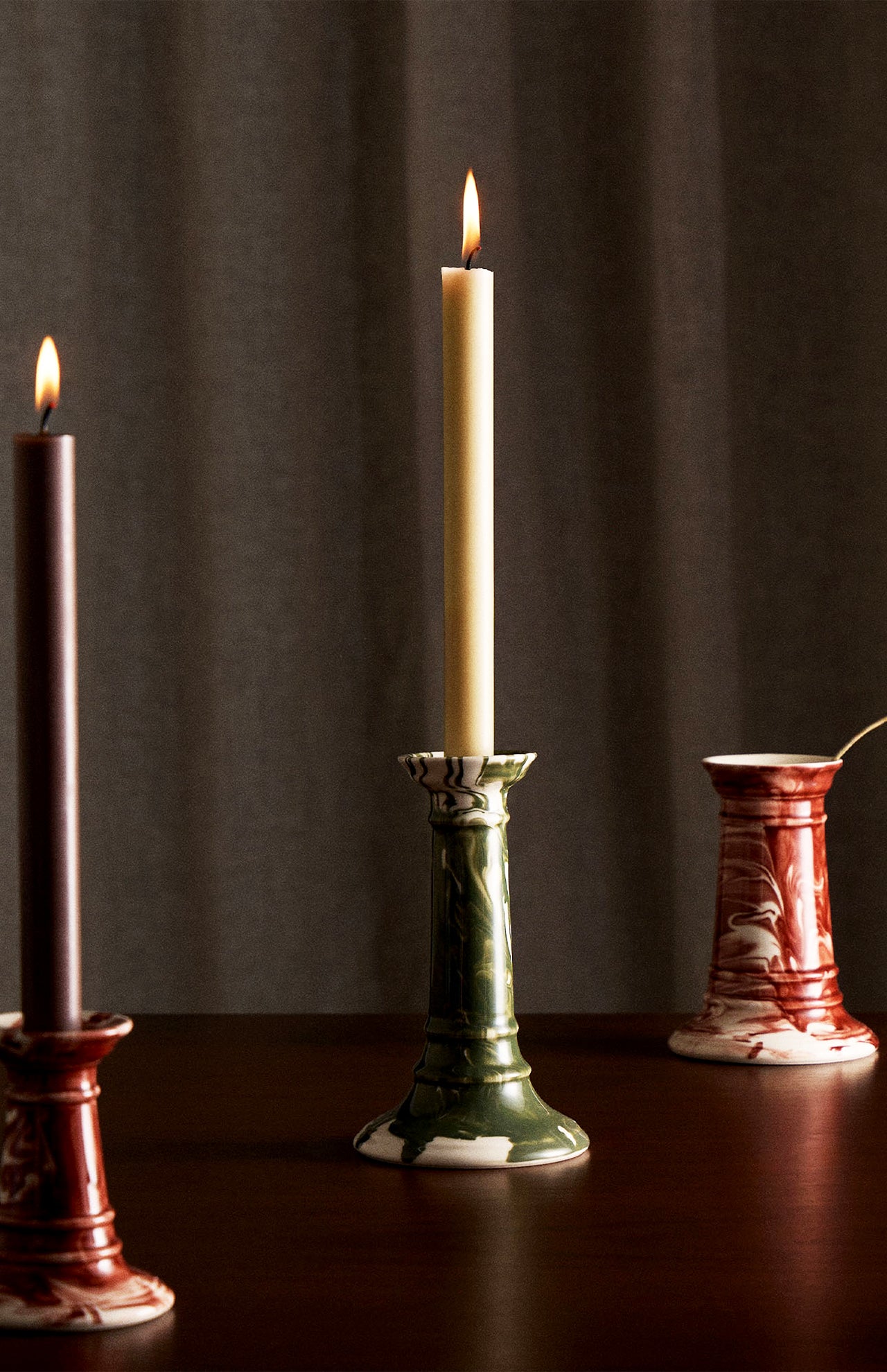 Three tall candles stand on a dark wooden surface. Two are lit—one beige in THE CONRAN SHOP’s Marbled Wardour Candlestick with hand-marbled glaze, and one dark brown in a red holder. A neutral, textured curtain forms the background.