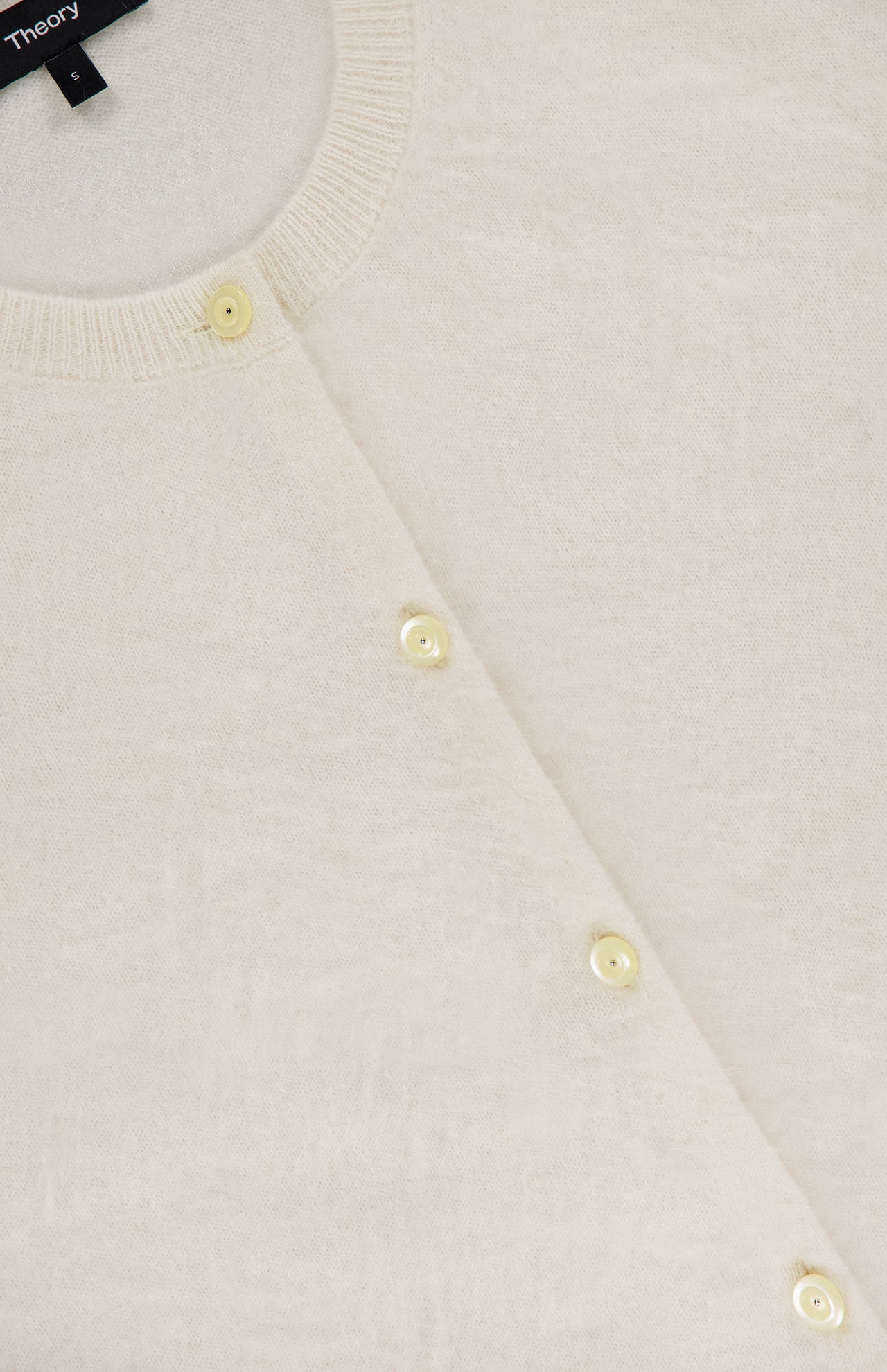 Close-up of THEORY's Brushed Cashmere Cardigan in ivory, featuring a round neckline, light buttons, and soft textured lightweight cashmere. A partially visible black brand label appears at the top left.