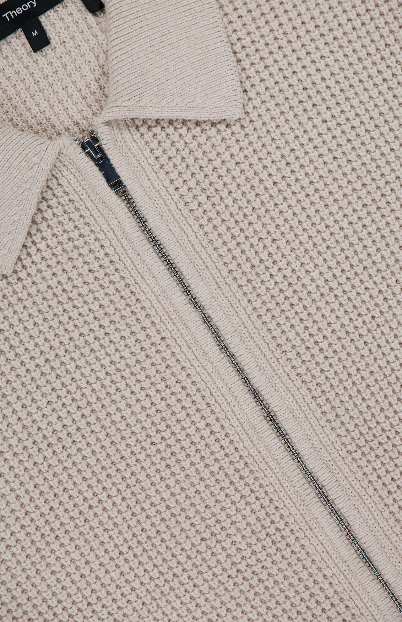 Close-up of THE­ORY's Gary Zip Sweater Jacket in beige waffle-knit cotton-cashmere, featuring a center zipper, folded collar, and a partially visible black label with white text at the top.