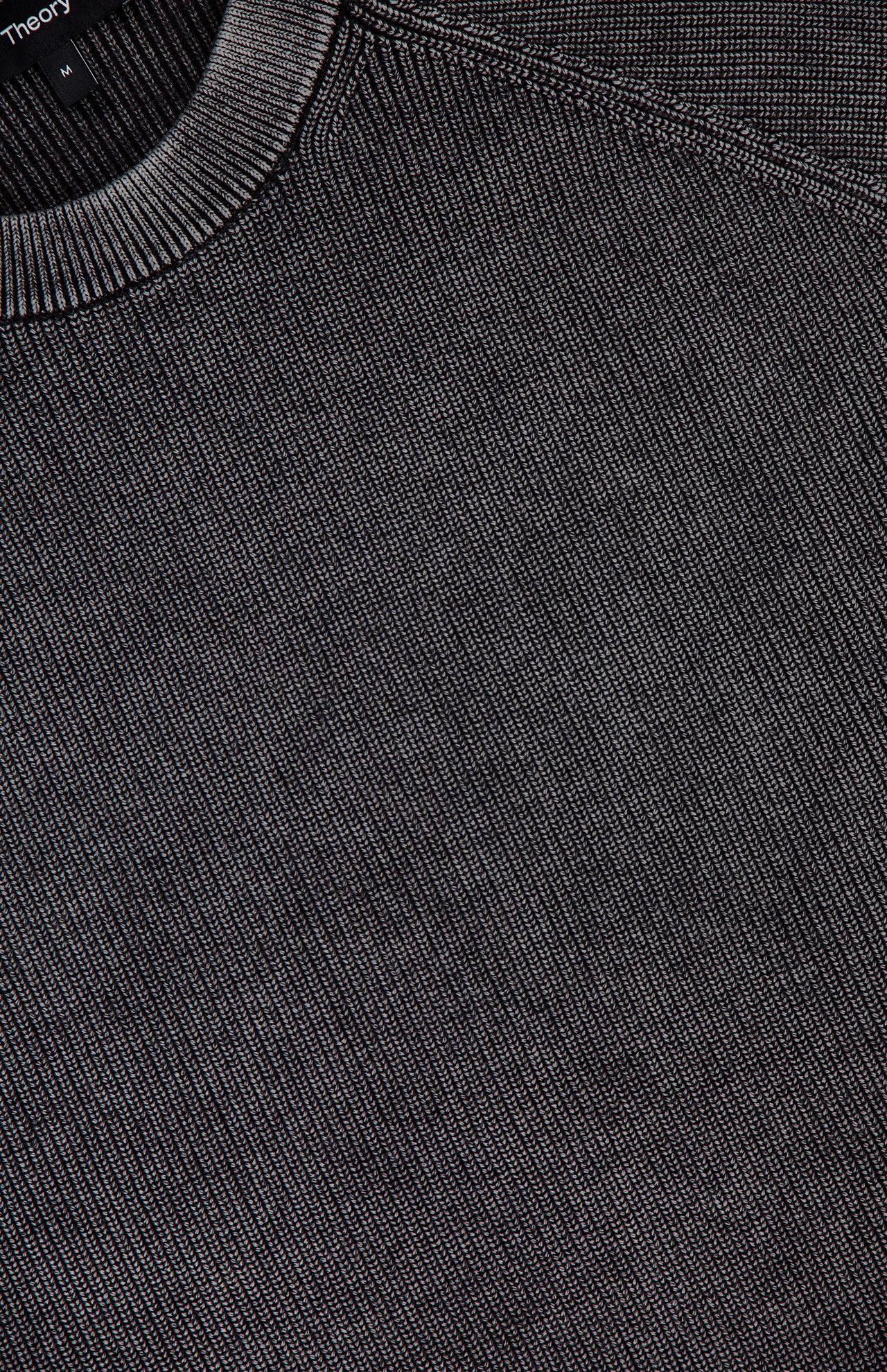Close-up of the THEORY Breach Wash Long Sleeve Crewneck Sweater in dark gray organic cotton, featuring ribbed knit texture, visible stitching at the neckline, and part of a black label near the top left corner.