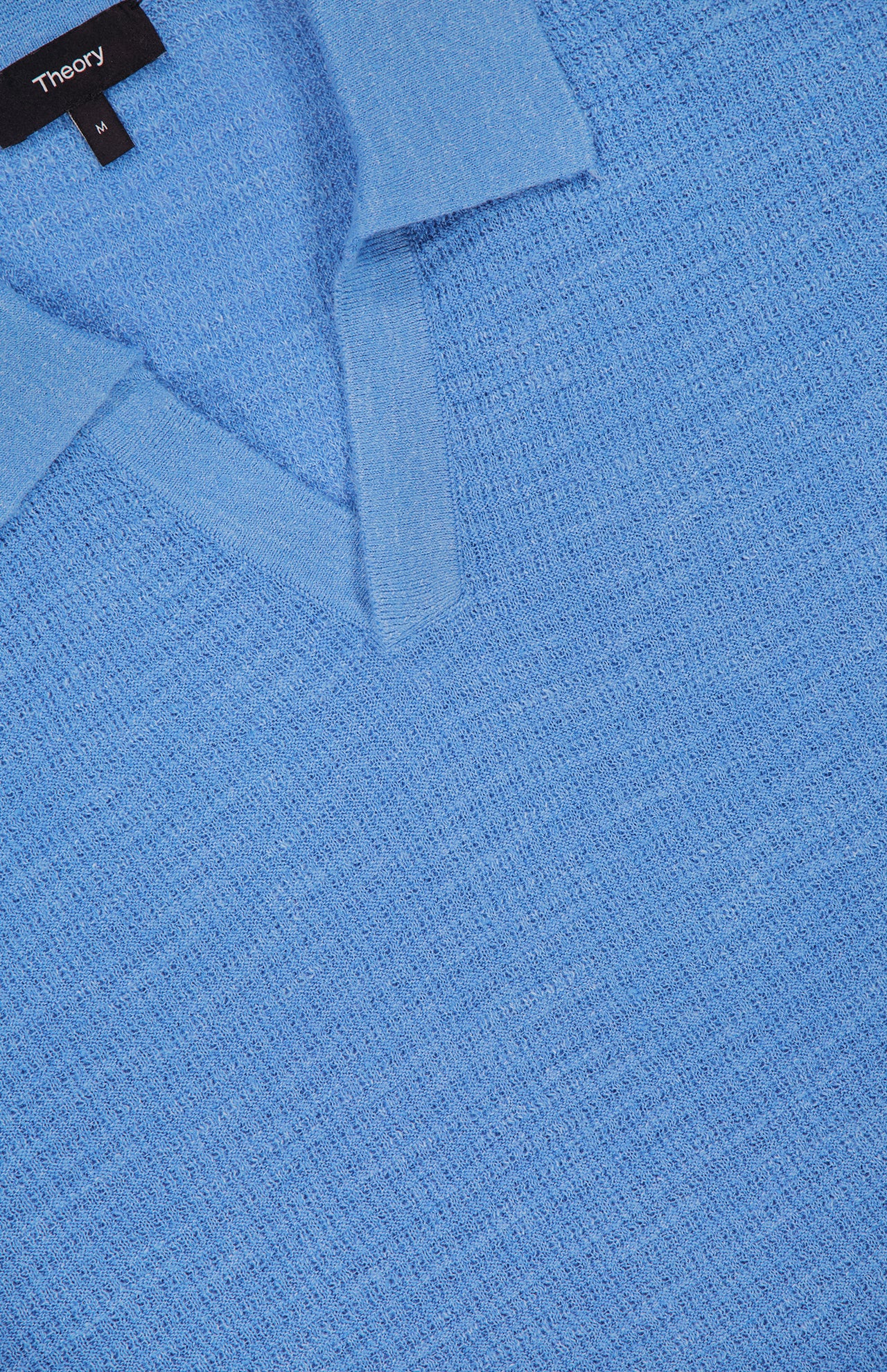 Close-up of the THEORY Brenan Knit Johnny Collar Polo Shirt in light blue, featuring a linen-blend knit with visible texture and a black THEORY label in size M.