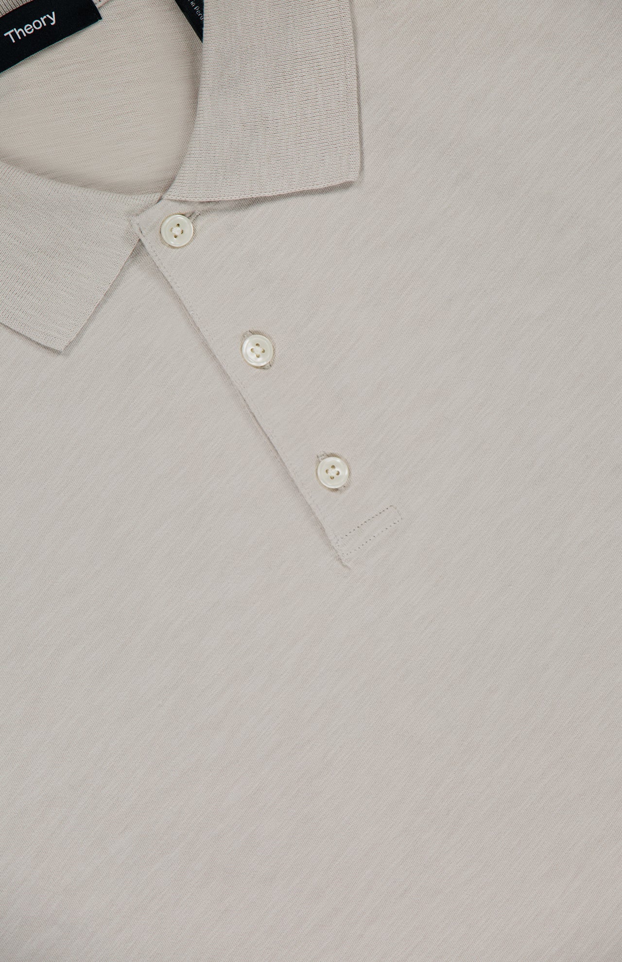 Close-up of the THEORY Bron Cosmos Polo Shirt in light beige, featuring three white buttons, a ribbed collar, and a partially visible Theory tag at the top.
