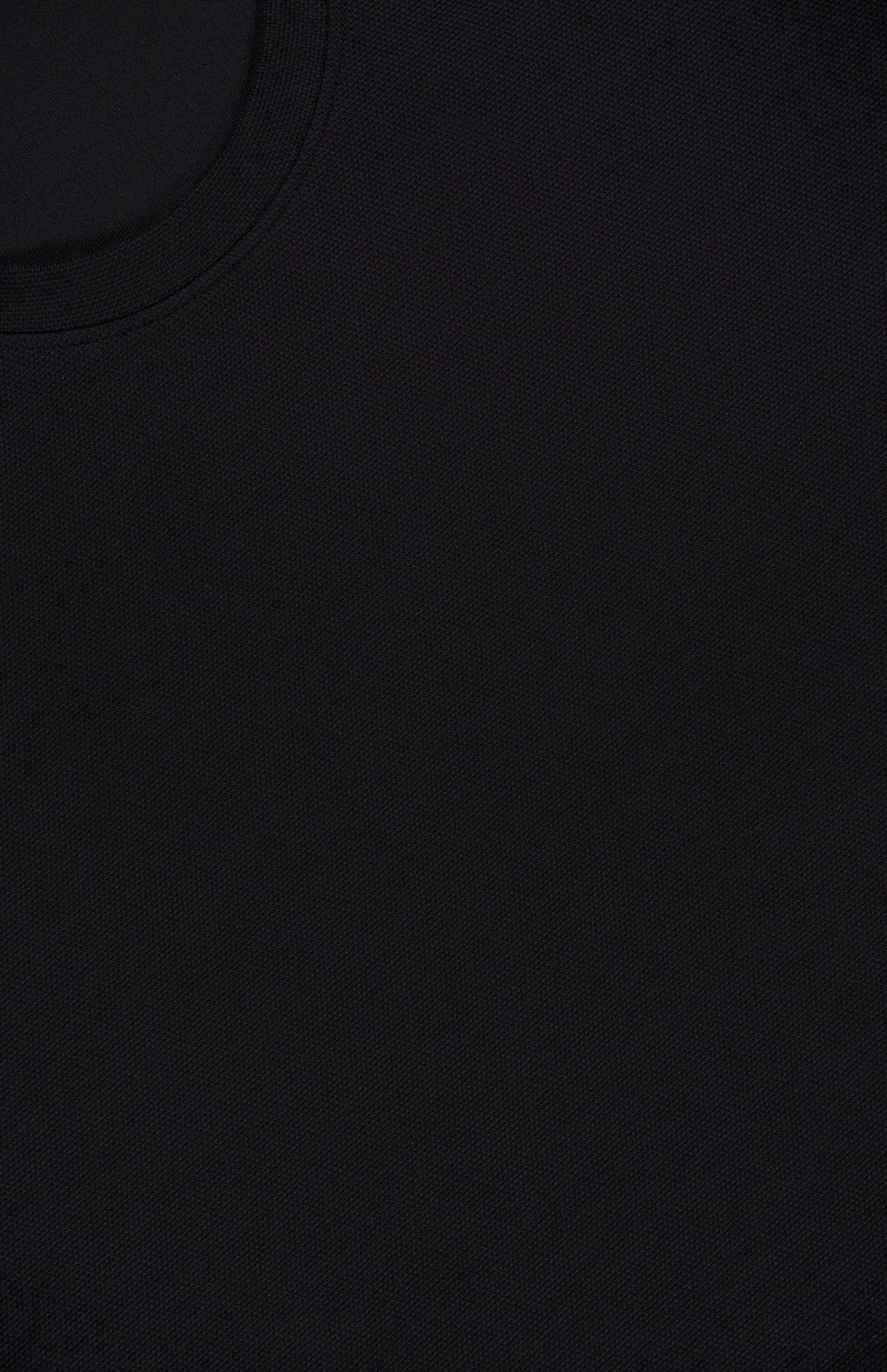 Close-up of the black textured fabric of the THEORY Delroy Cotton-blend T-Shirt, featuring a visible curved seam or collar in the upper left corner. The material is a double pique jersey, commonly chosen for its subtle texture and stretch—ideal for premium black t-shirts like this one.
