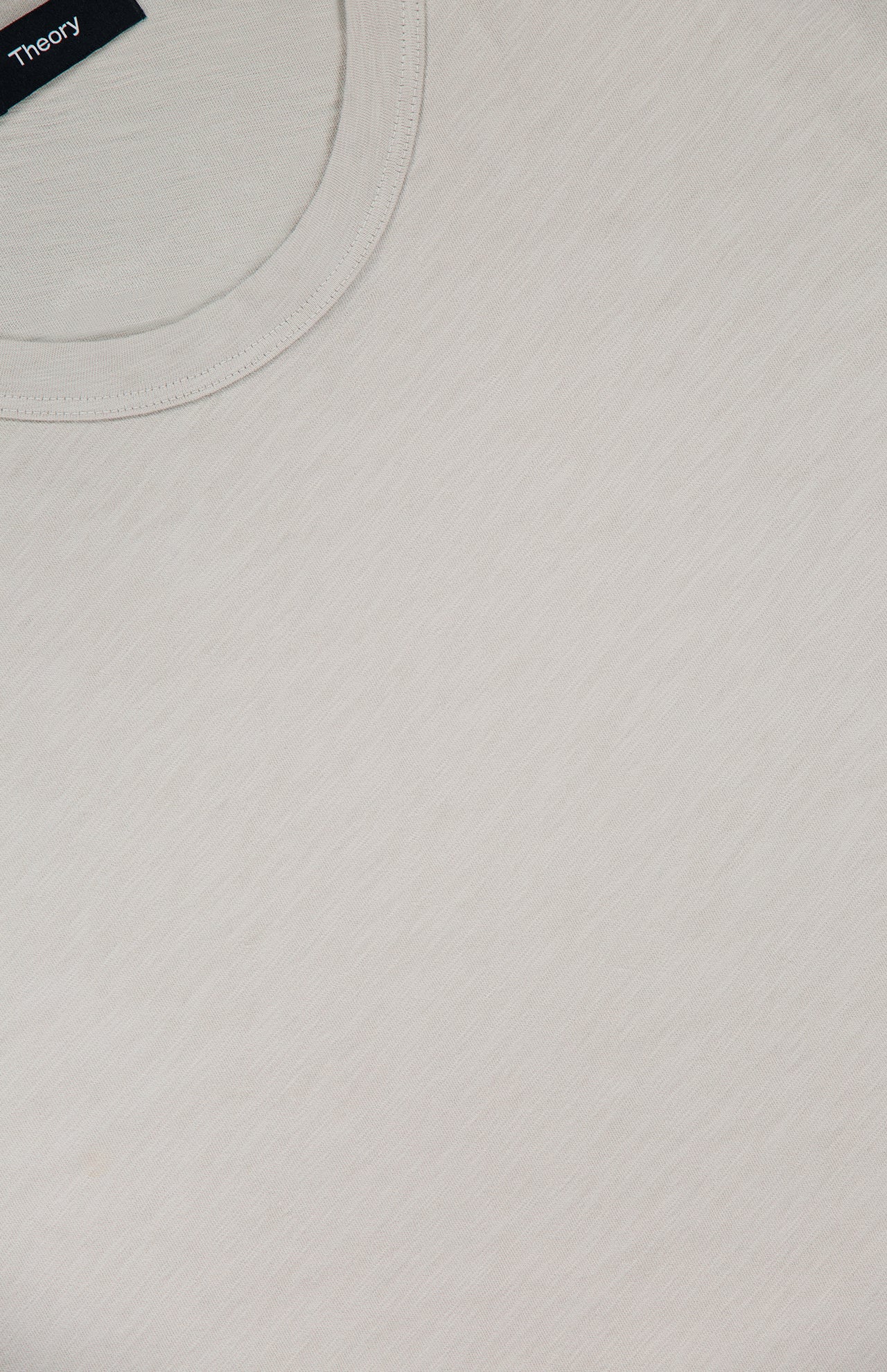 Close-up of the Essential Cosmo T-shirt by THEORY—a plain, lightweight light grey or off-white jersey tee with a round neckline and a visible black THEORY label in the top left corner. Made in Peru.
