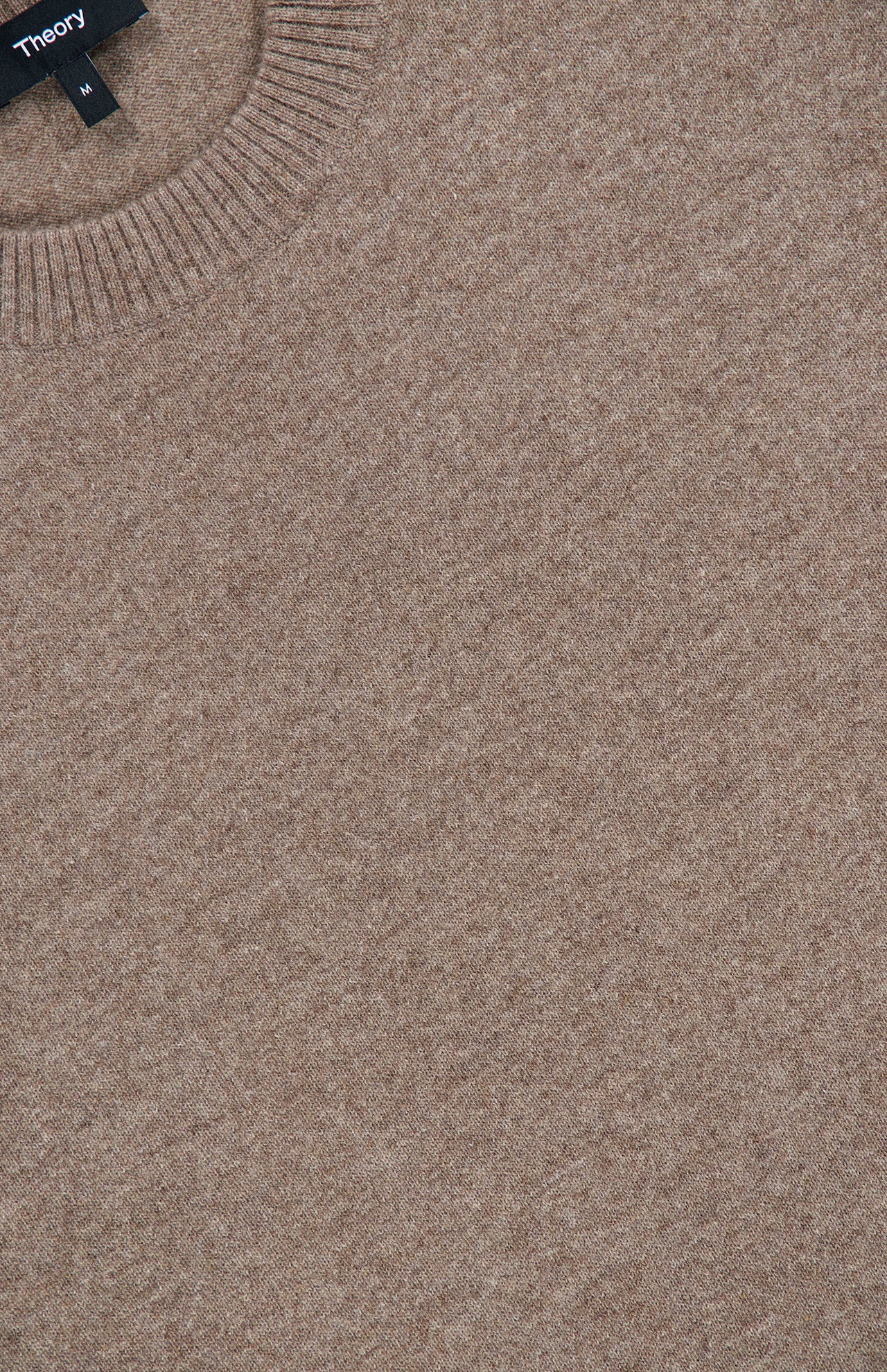 Close-up of the Hilles Cashmere Crewneck Sweater in light brown, featuring a minimalist knit design with a ribbed crew neckline. A black THEORY brand tag displaying the size M is visible at the top left of the image, emphasizing this classic sweater’s refined simplicity.