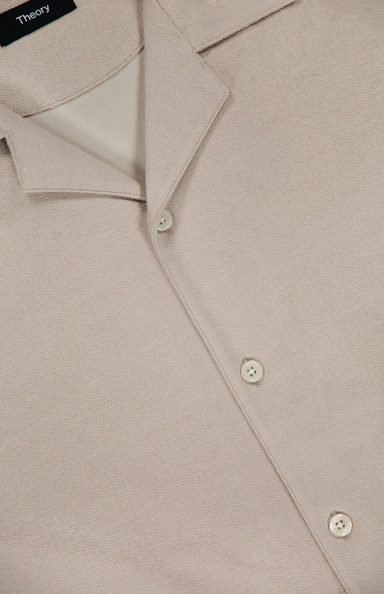 Close-up of the Soft Terry Short Sleeve Shirt by THEORY, in beige with a textured finish, pointed collar, and three white buttons. This casual work shirt exemplifies refined style and effortless sophistication.