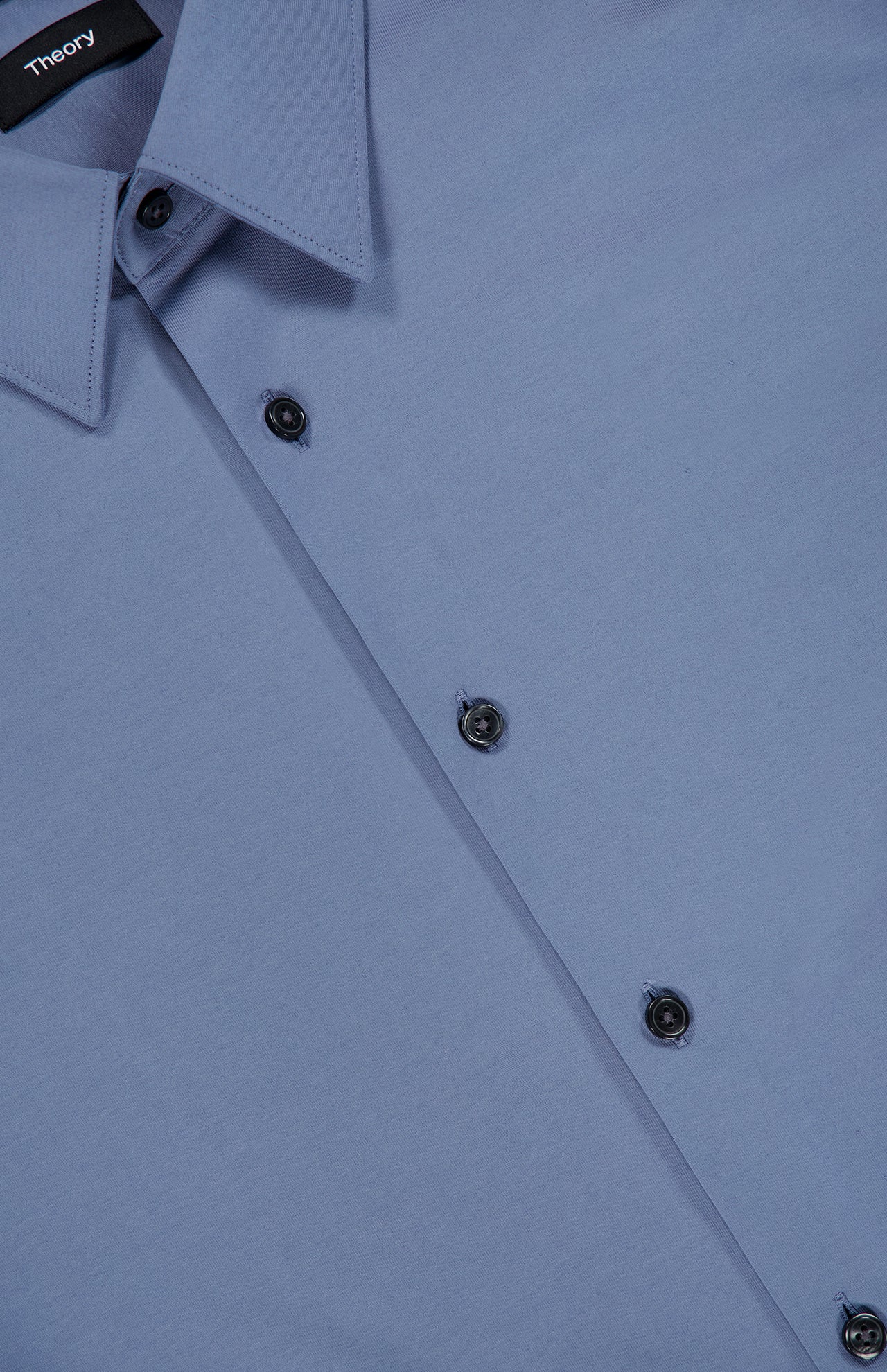 Close-up of a neatly buttoned, slim fit light blue Sylvain Structure Dress Shirt with black buttons and a pointed collar. The THEORY brand label is visible inside the collar, emphasizing the shirt’s modern silhouette and wrinkle-resistant fabric.