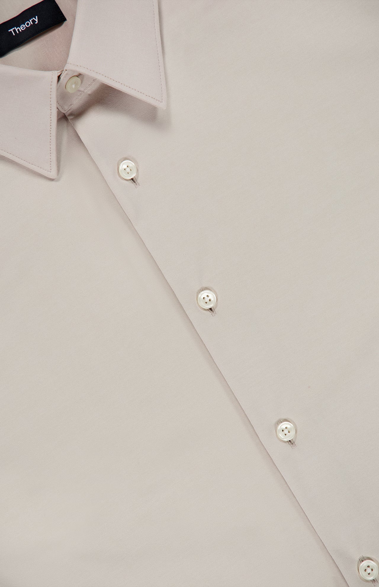 A close-up of the Sylvain Structure Dress Shirt by THEORY in light beige showcases its pointed collar, four white buttons, and a prominent black tag featuring the THEORY logo in white text. This dress shirt is wrinkle-resistant, machine-washable, and expertly blends classic design with easy care.