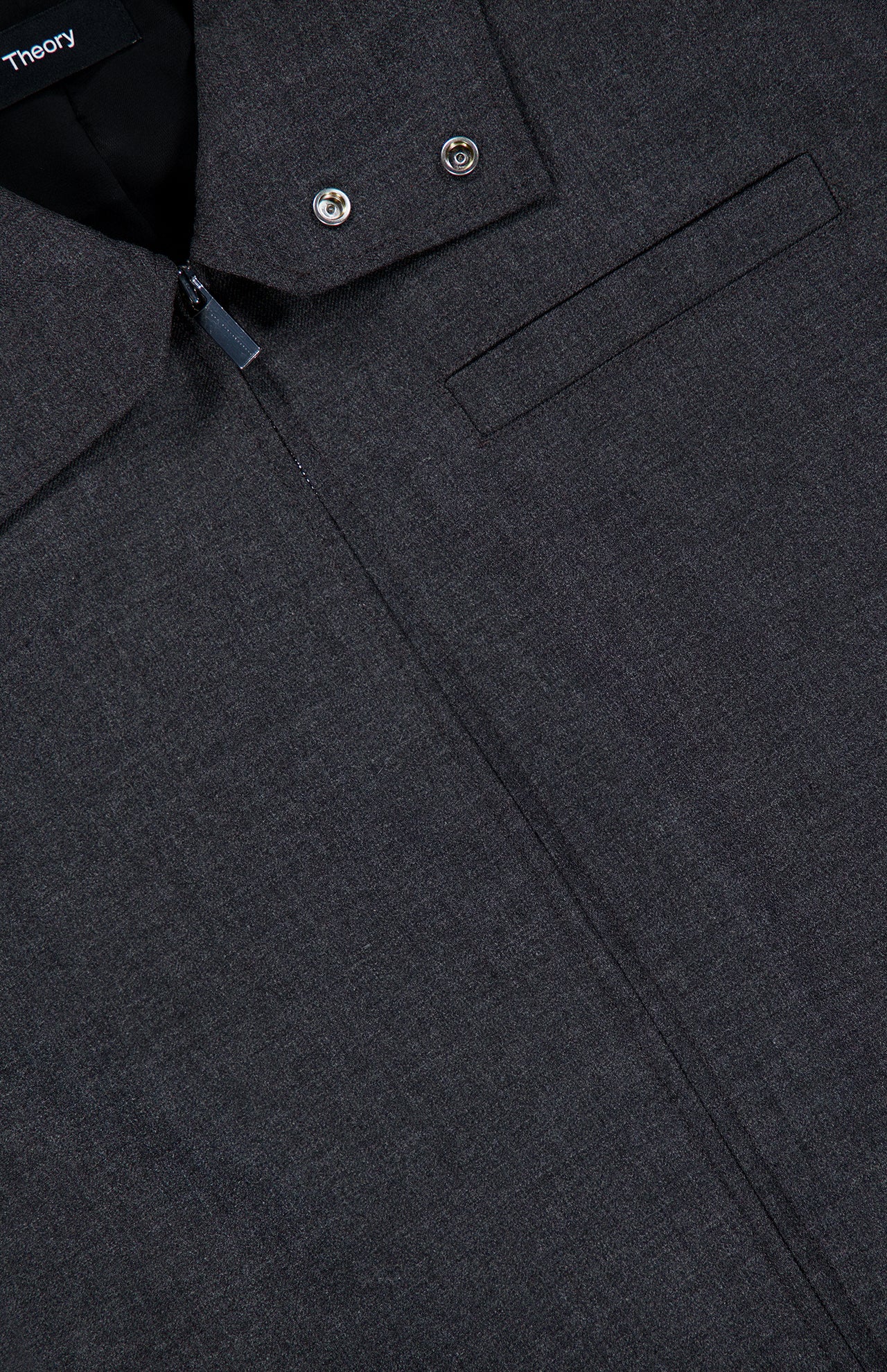 Close-up of the Harrington Cropped Jacket by THEORY in dark gray charcoal. This stylish wool-blend piece features a zip-up front, chest pocket, and two metal buttons near the collar. The visible fabric texture and part of the THEORY brand label highlight its sophisticated design—making it perfect as an office jacket or for elevating everyday outfits.