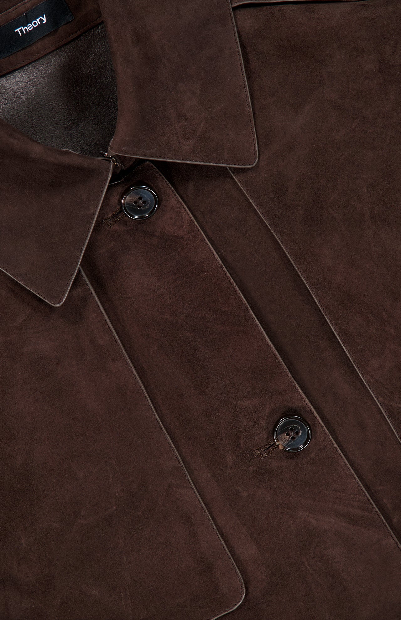 Close-up of the Reece Cropped Trench Coat by THEORY in chocolate brown suede, featuring large lapels, two dark buttons, and visible stitching. A small black THEORY tag is partially visible at the top left.