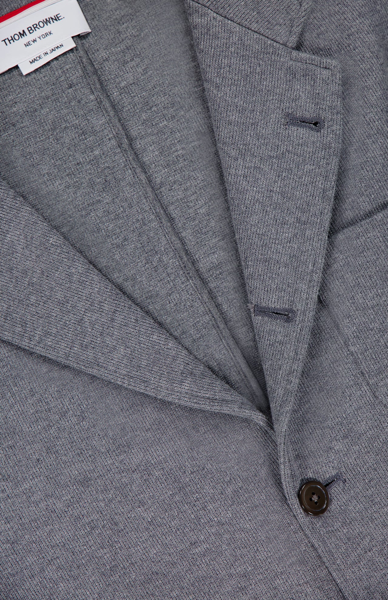 Close-up of the THOM BROWNE Cotton Knit Sack Jacket in grey, featuring a notched lapel, single dark button, and an interior label that reads "Thom Browne New York Made in Japan," highlighting this refined men’s blazer.