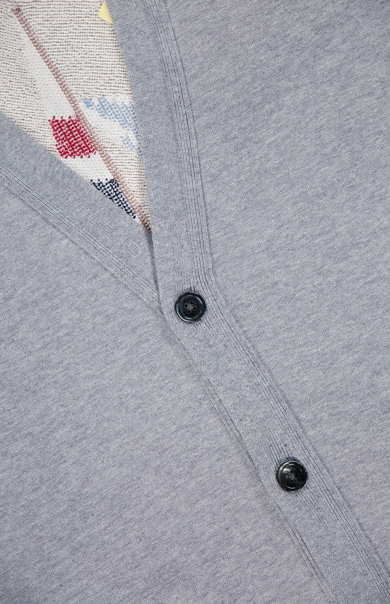 Close-up of THOM BROWNE’s Stitched Boat Graphic Cardigan in light grey, styled as a men’s Americana piece with two black buttons. A patterned knit top in red, blue, and beige is visible under the cardigan opening.