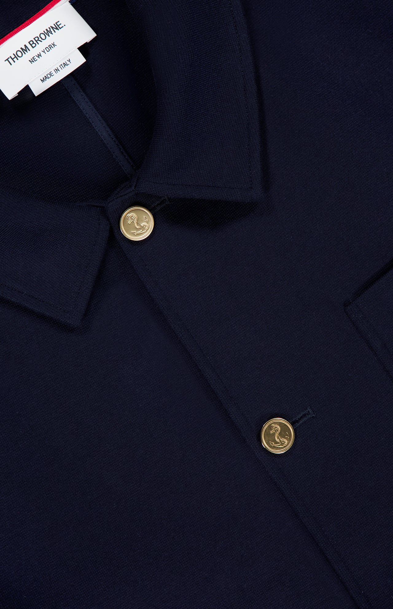 Close-up of a dark navy Compact Milano Car Jacket by THOM BROWNE, featuring gold engraved buttons, a pointed collar, and the brand’s label at the neckline—an elevated take on classic Americana style.