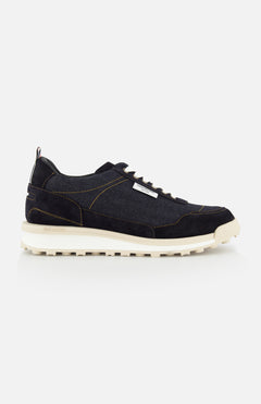 The THOM BROWNE Denim Alumni Trainer Sneaker for men features a navy blue design with white laces, a beige sole, and yellow stitching, showcased in a side profile against a plain white background.