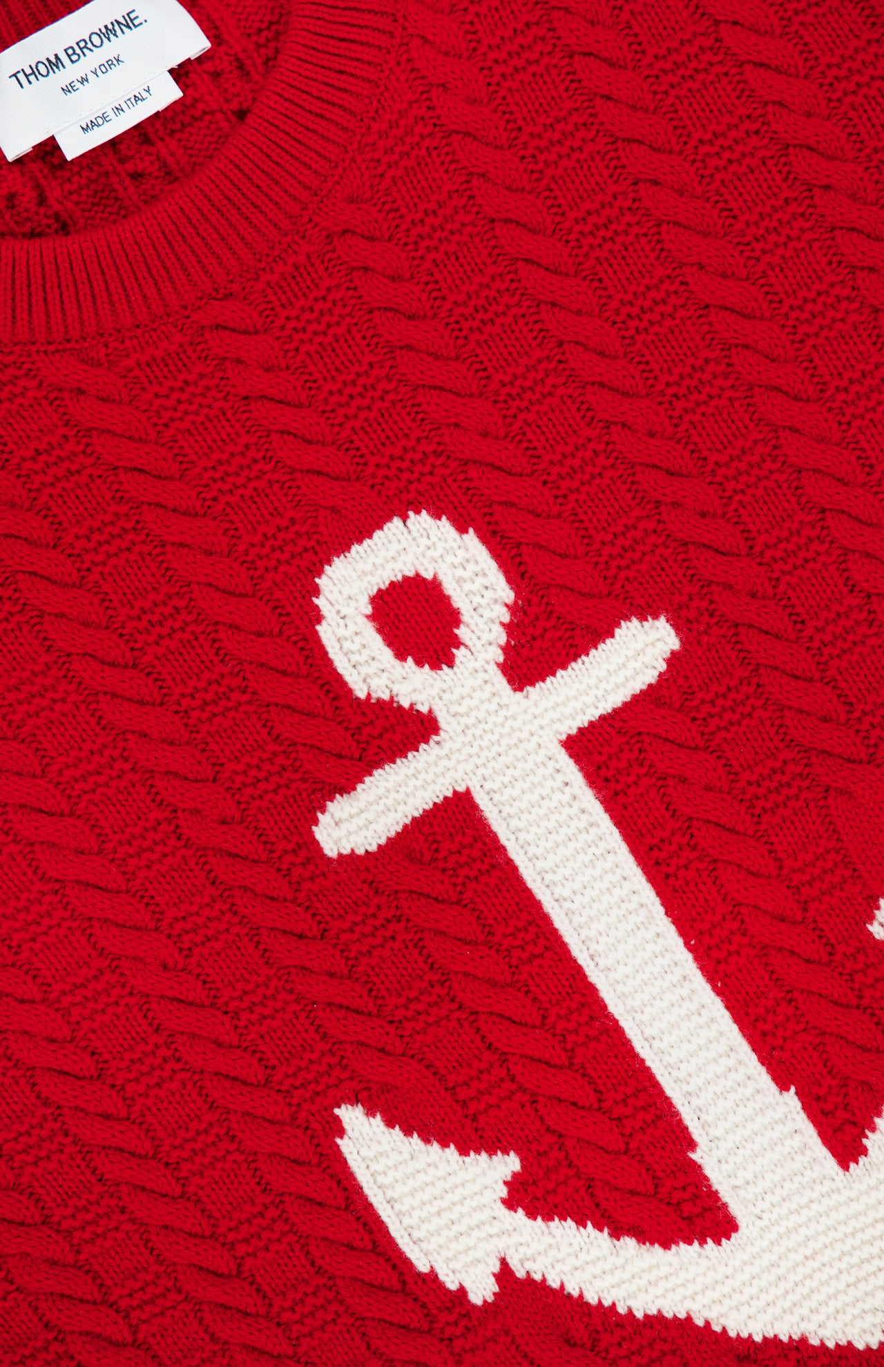 Introducing the Intarsia Anchor Merino Wool Crew Neck Sweater by THOM BROWNE—a vibrant red cable-knit sweater crafted from fine merino wool, featuring a bold white anchor intarsia on the front for a signature nautical flair. A label at the neckline reads “THOM BROWNE. NEW YORK. MADE IN ITALY.”.