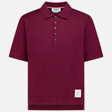 The THOM BROWNE Jersey Short Sleeve Rib Cuff Polo Shirt, a men's classic in burgundy, showcases a smart collar, five-button placket, ribbed cuffs, and a signature small white label near the bottom hem. Presented against a plain white background.