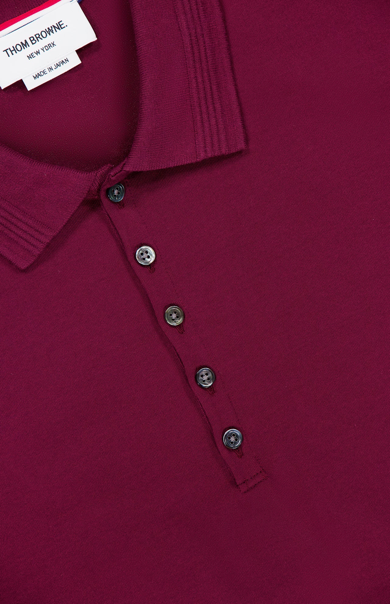 Close-up of the THOM BROWNE Jersey Short Sleeve Rib Cuff Polo Shirt in rich burgundy, detailed with five black buttons and a visible white collar tag reading “THOM BROWNE. NEW YORK. MADE IN JAPAN”—a refined nod to classic Americana style.