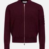 The THOM BROWNE Milano Stitch Zip Up Bomber Jacket is a burgundy jacket crafted in the classic Americana zip-up style, featuring ribbed cuffs, practical pockets, and a distinctive horizontal stitching detail on one sleeve. It is presented against a plain white background.