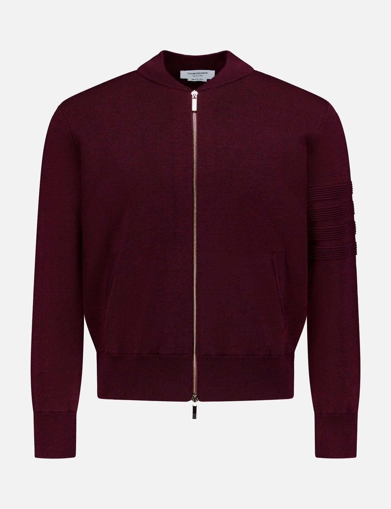 The THOM BROWNE Milano Stitch Zip Up Bomber Jacket is a burgundy jacket crafted in the classic Americana zip-up style, featuring ribbed cuffs, practical pockets, and a distinctive horizontal stitching detail on one sleeve. It is presented against a plain white background.