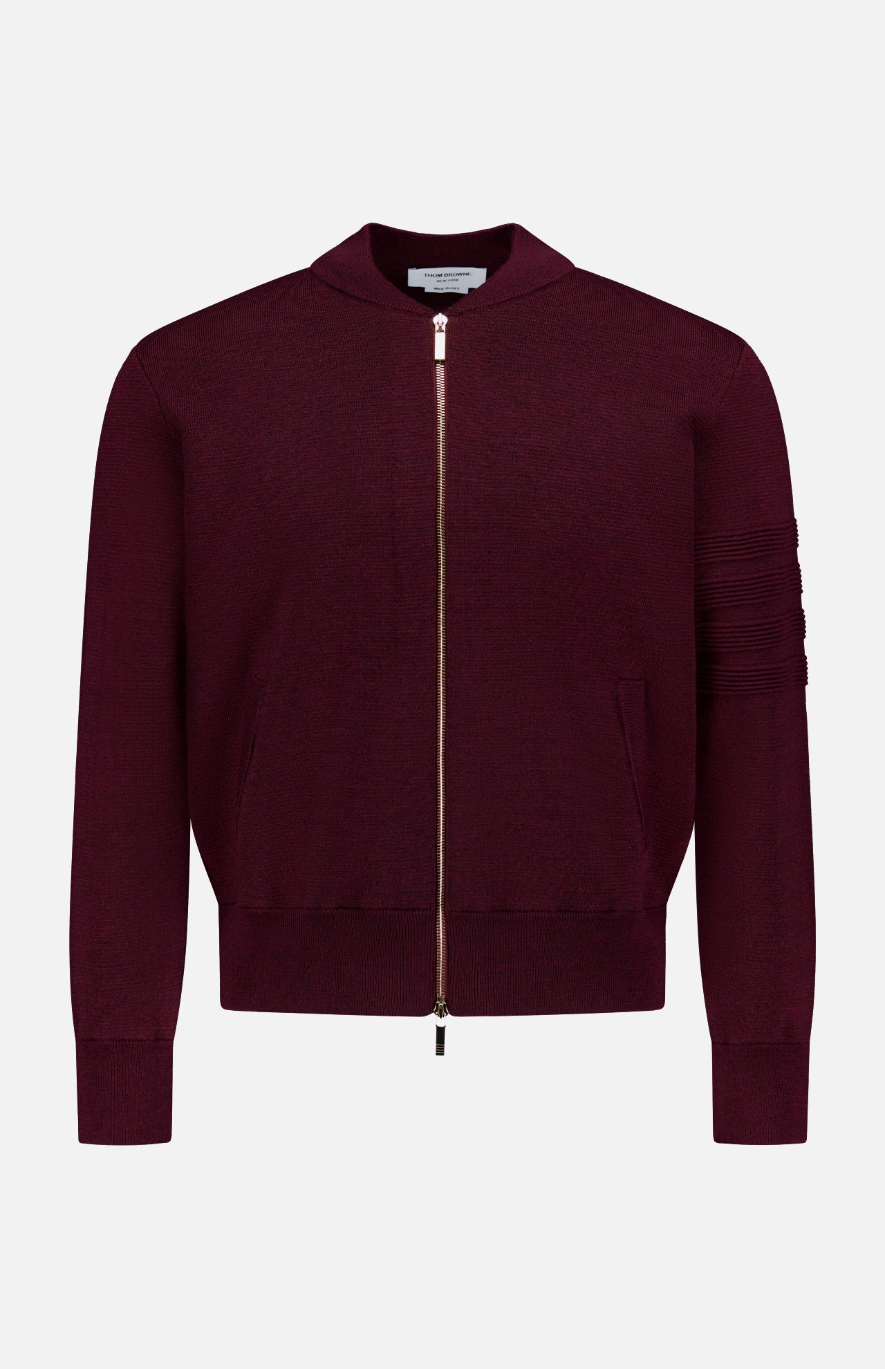 The THOM BROWNE Milano Stitch Zip Up Bomber Jacket is a burgundy jacket crafted in the classic Americana zip-up style, featuring ribbed cuffs, practical pockets, and a distinctive horizontal stitching detail on one sleeve. It is presented against a plain white background.