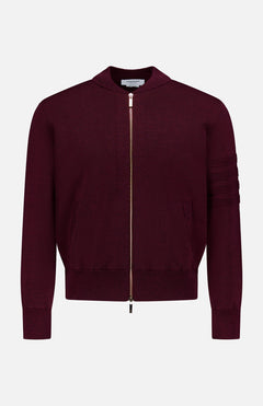 The THOM BROWNE Milano Stitch Zip Up Bomber Jacket is a burgundy jacket crafted in the classic Americana zip-up style, featuring ribbed cuffs, practical pockets, and a distinctive horizontal stitching detail on one sleeve. It is presented against a plain white background.