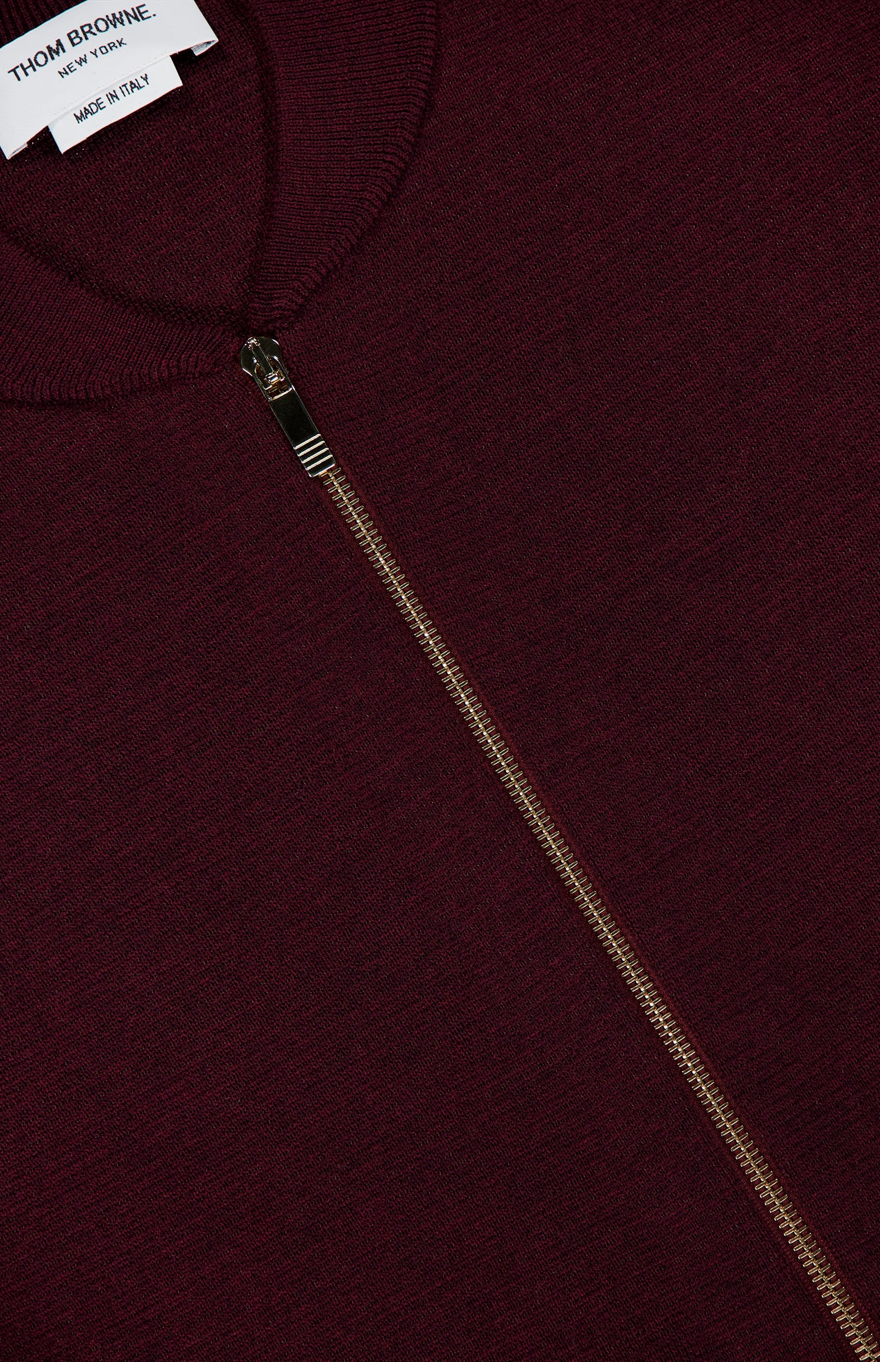 Close-up of a burgundy Milano Stitch Zip Up Bomber Jacket featuring a silver zipper. A white clothing label in the top left corner reads THOM BROWNE. NEW YORK. MADE IN ITALY.