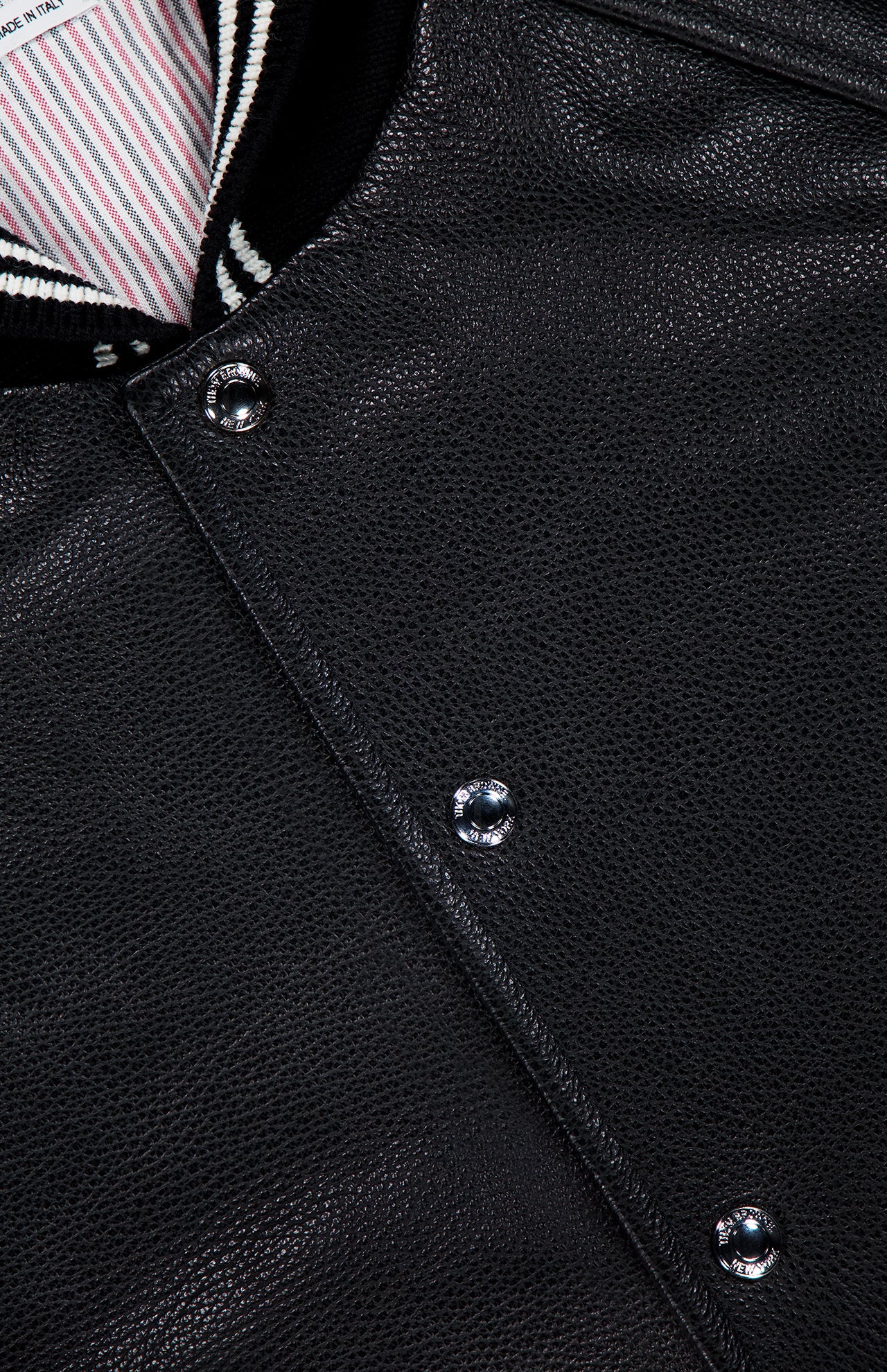 Close-up of the THOM BROWNE Oversized Deerskin Leather Bomber Jacket, with textured black leather, silver snap buttons, and a black-and-white striped ribbed collar for understated Americana style.