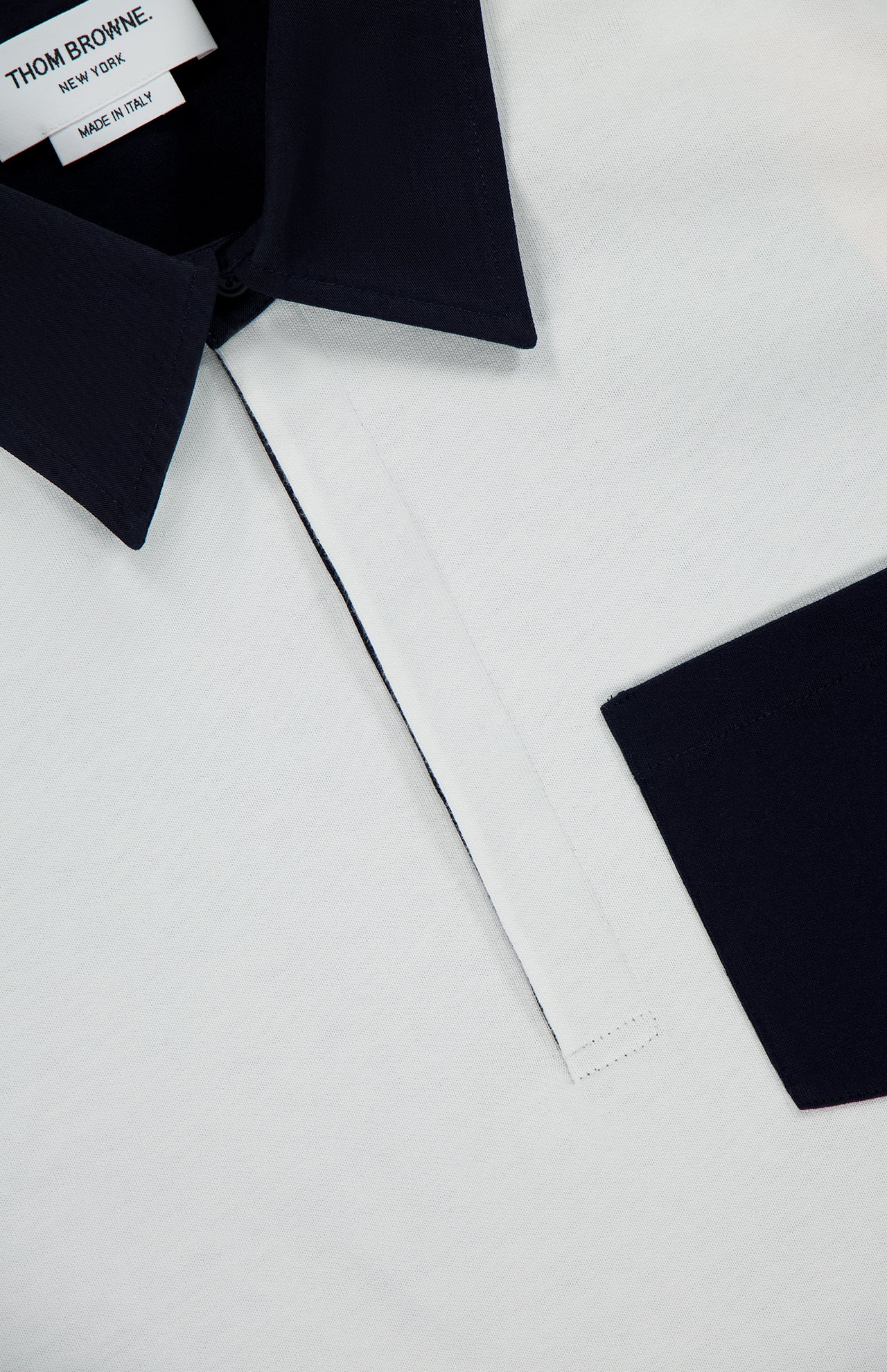 Close-up of the THOM BROWNE Oversized Long Sleeve Colorblock Rugby Polo Shirt, showcasing a white body contrasted by a dark navy collar and sleeve cuffs in an elegant colorblock design. The visible label reads “THOM BROWNE. NEW YORK. MADE IN ITALY,” and the placket is neatly stitched in white.