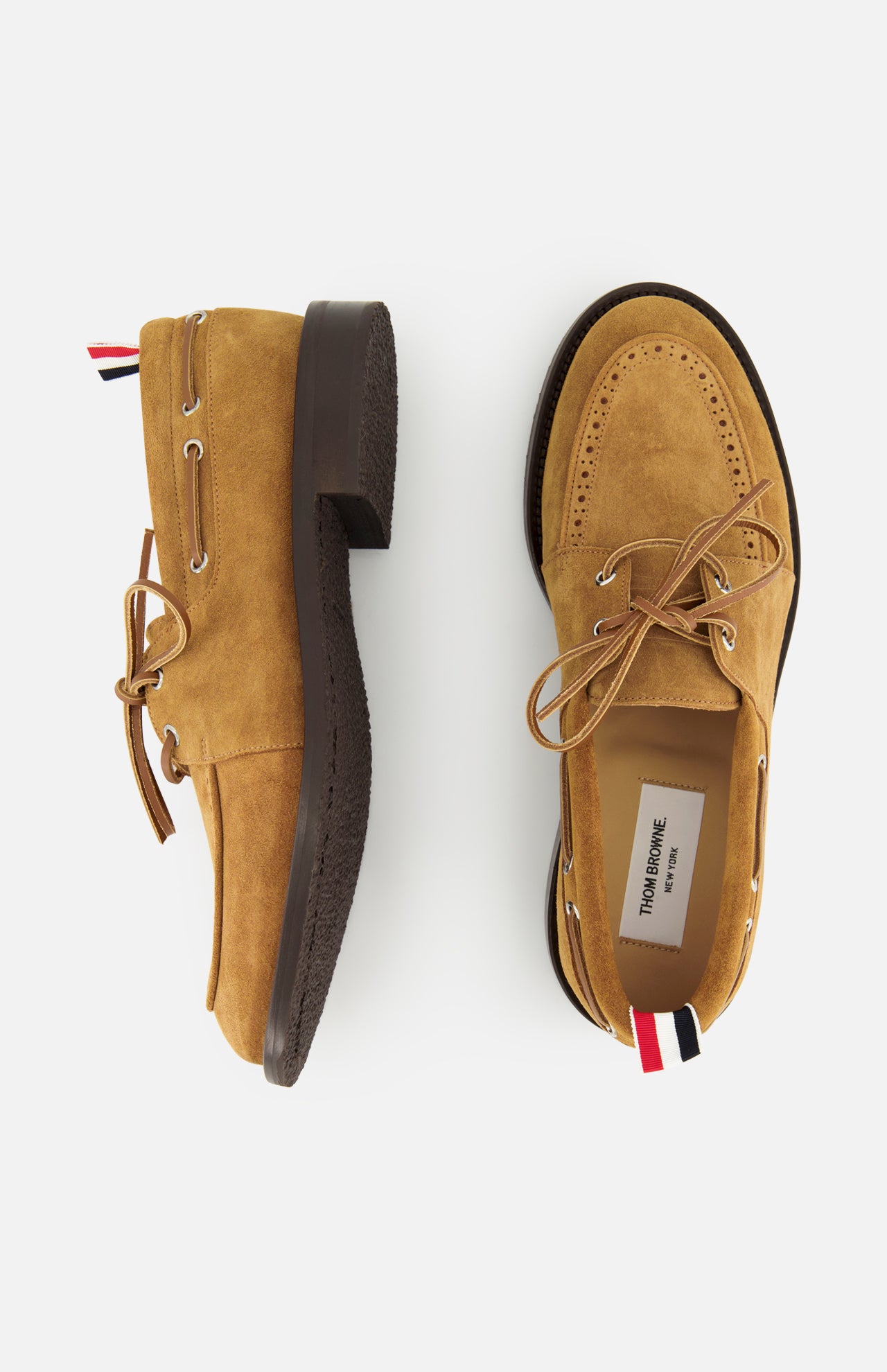 The THOM BROWNE Padded Boat Suede Loafers in tan feature brown soles, beige laces, and signature red, white, and blue striped heel tabs. Displayed against a white background, one is shown from the side and one from above.