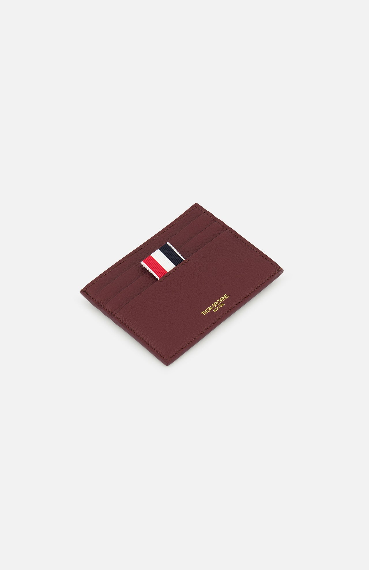 The THOM BROWNE Pebble Grain Leather Card Holder features four card slots, gold embossed details, and a red, white, and navy striped fabric tab. Shown on a white background for a refined look.
