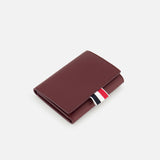 The THOM BROWNE Pebble Grain Leather Trifold Wallet, in closed burgundy leather with a textured finish and a red, white, and black fabric tab, is featured against a plain white background.