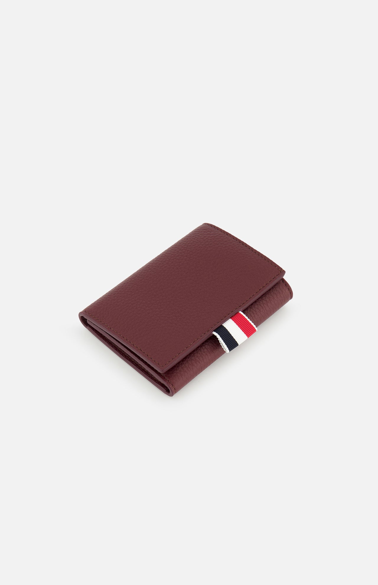 The THOM BROWNE Pebble Grain Leather Trifold Wallet, in closed burgundy leather with a textured finish and a red, white, and black fabric tab, is featured against a plain white background.