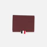 The THOM BROWNE Pebble Grain Leather Trifold Wallet in burgundy is shown closed on a plain white background, featuring a distinctive red, white, and black fabric tab at the bottom.