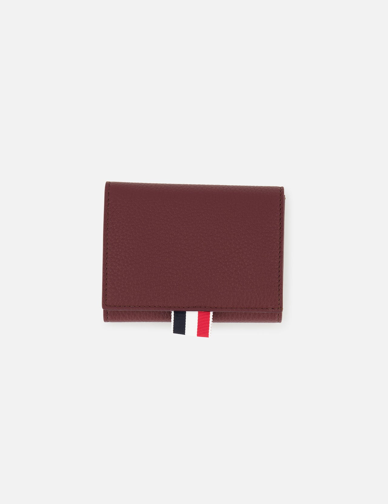 The THOM BROWNE Pebble Grain Leather Trifold Wallet in burgundy is shown closed on a plain white background, featuring a distinctive red, white, and black fabric tab at the bottom.