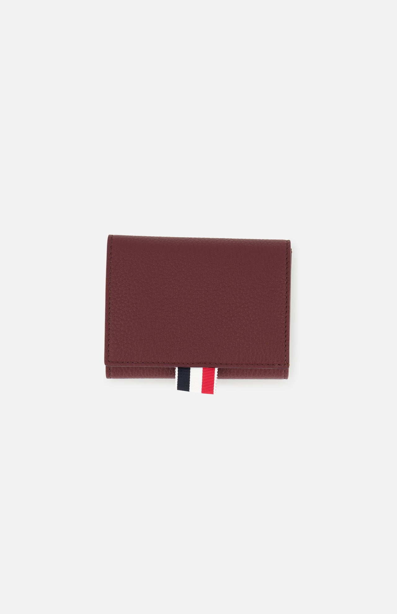 The THOM BROWNE Pebble Grain Leather Trifold Wallet in burgundy is shown closed on a plain white background, featuring a distinctive red, white, and black fabric tab at the bottom.