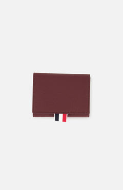 The THOM BROWNE Pebble Grain Leather Trifold Wallet in burgundy is shown closed on a plain white background, featuring a distinctive red, white, and black fabric tab at the bottom.