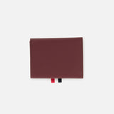 The THOM BROWNE Pebble Grain Leather Trifold Wallet is shown closed on a white background, featuring a textured burgundy finish and two ribbon tabs—one red and one black—extending from the bottom edge.