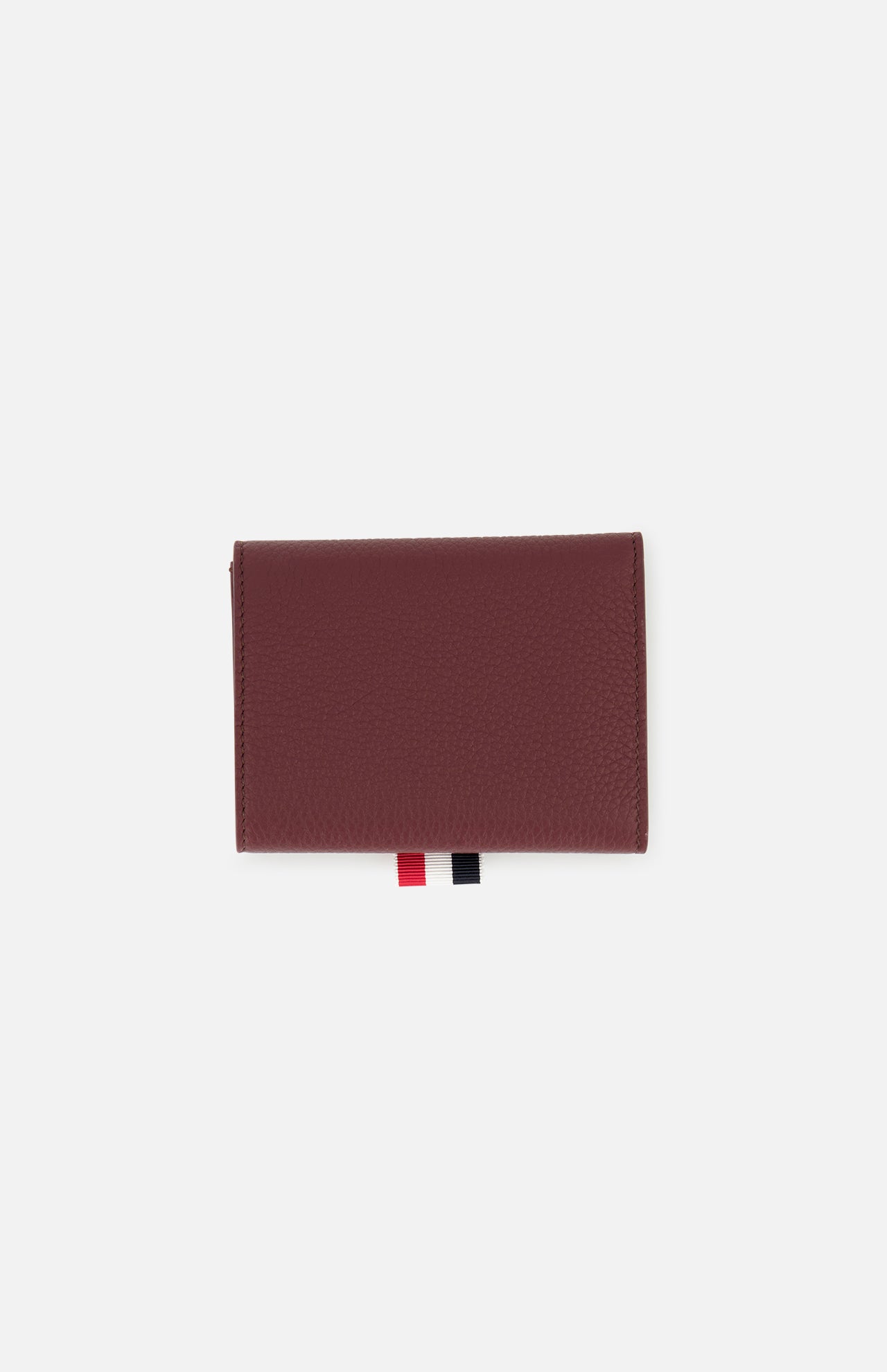 The THOM BROWNE Pebble Grain Leather Trifold Wallet is shown closed on a white background, featuring a textured burgundy finish and two ribbon tabs—one red and one black—extending from the bottom edge.