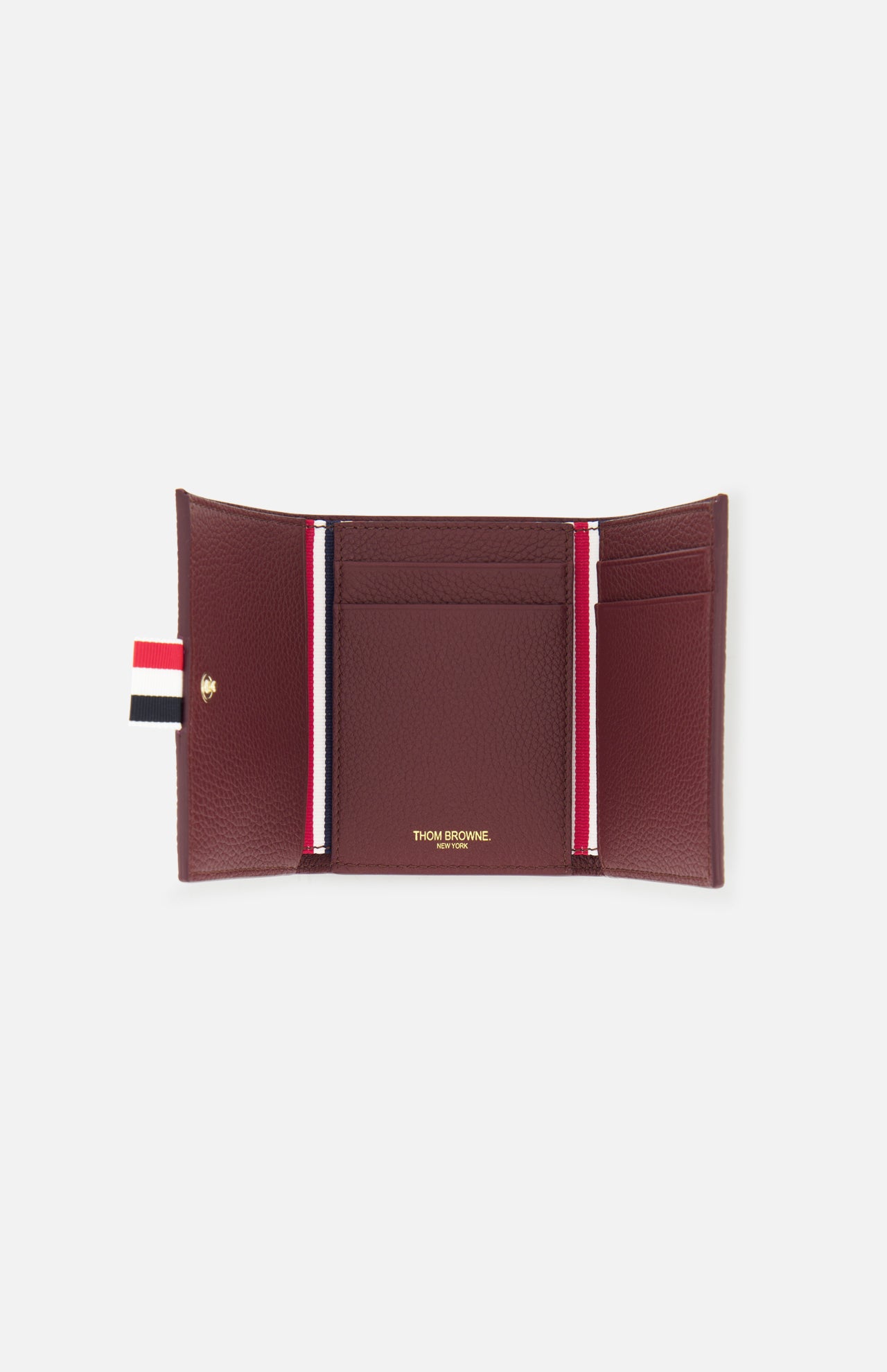The Pebble Grain Leather Trifold Wallet by THOM BROWNE features burgundy leather, gold branding inside, two card slots per side, a central pocket, and a red, white, and blue striped tab—a refined men’s accessory for everyday style.