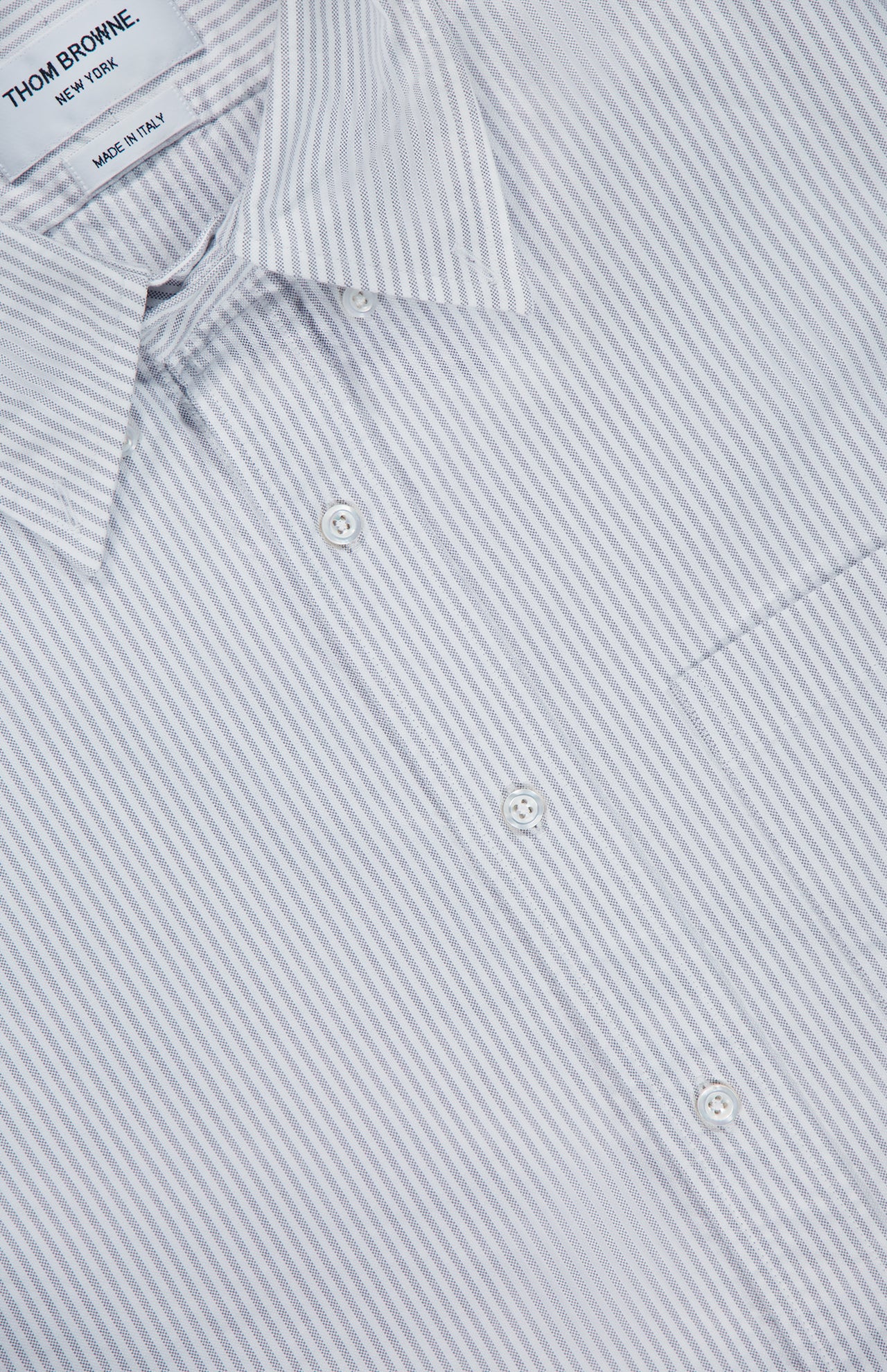 Close-up of the THOM BROWNE Straight Fit Lobsters Long Sleeve Oxford Shirt, showcasing its white fabric with thin gray vertical stripes, a pointed collar, and part of a chest pocket. The label inside the collar reads THOM BROWNE. NEW YORK. MADE IN ITALY.