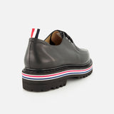 The THOM BROWNE Vitello Calf Leather Derby Loafer features black leather, a moc toe, chunky lug sole, red-white-blue midsole stripes, and a matching heel pull tab. Shown from the rear side on a white background.