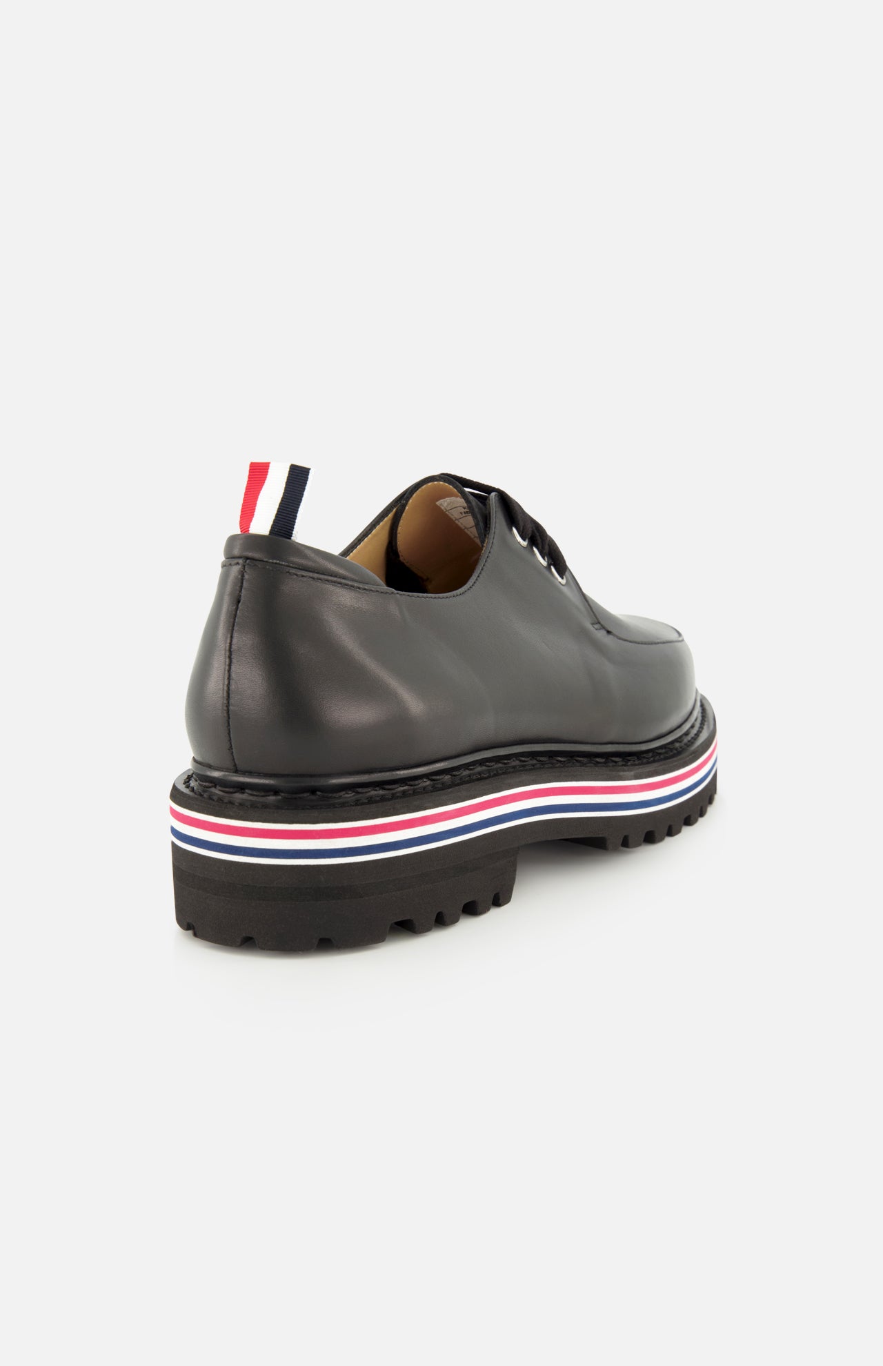 The THOM BROWNE Vitello Calf Leather Derby Loafer features black leather, a moc toe, chunky lug sole, red-white-blue midsole stripes, and a matching heel pull tab. Shown from the rear side on a white background.