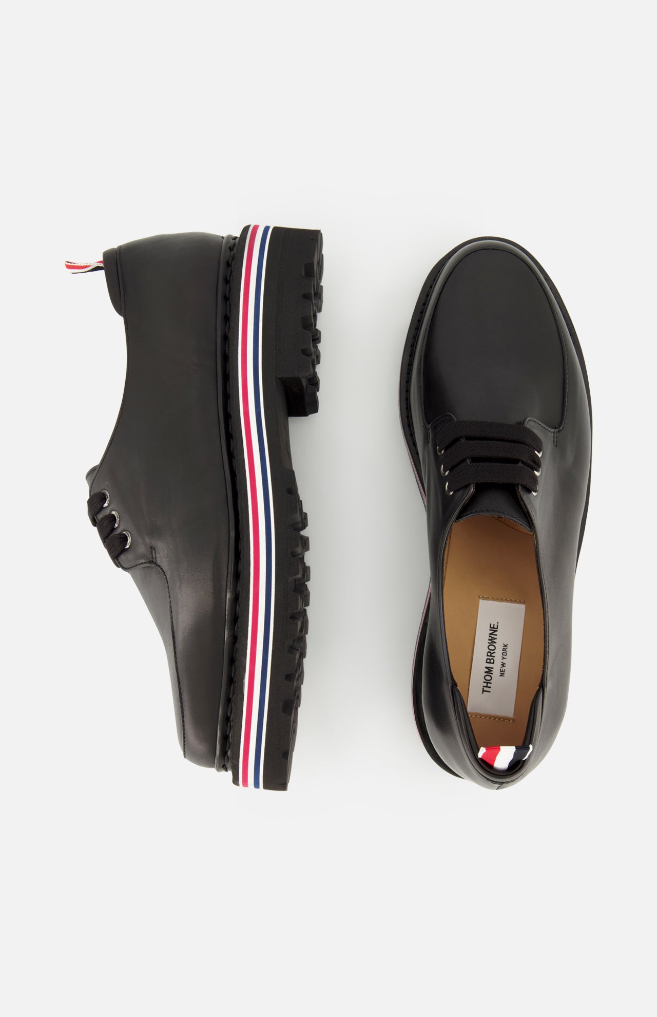 Two THOM BROWNE Vitello Calf Leather Derby Loafers in black are shown on a white background—one side view, one top view. The thick rubber soles display a red, white, and blue stripe along the side.