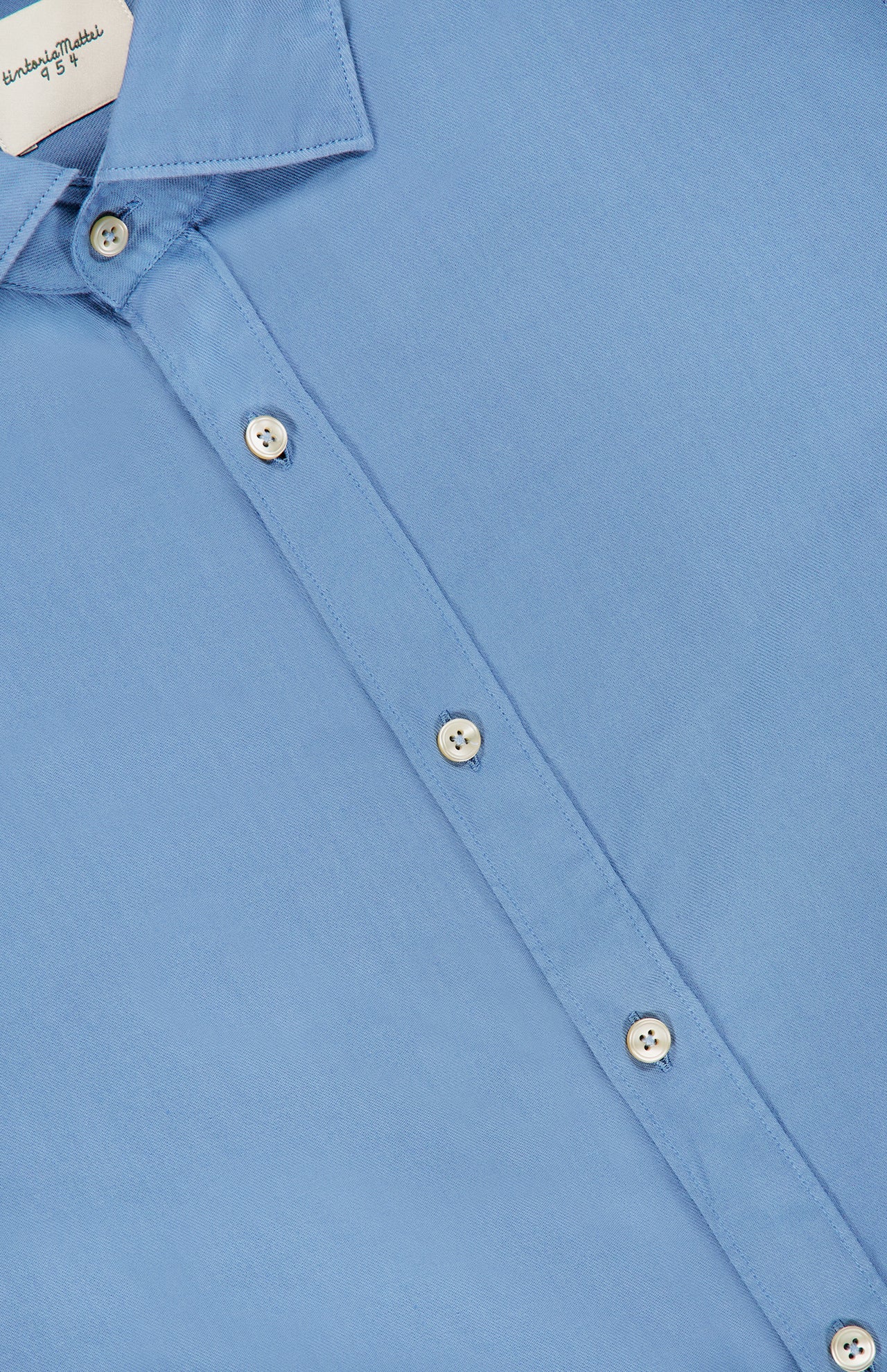 Close-up of the TINTORIA MATTEI Cotton Long Sleeve Dress Shirt in light blue, featuring white buttons and a pointed collar, laid flat. Part of the shirt’s label is visible inside the collar.