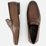 The Aldrin Leather Penny Loafer by TO BOOT is shown in side and top views on a white background, featuring brown polished leather with classic stitching around the toe.
