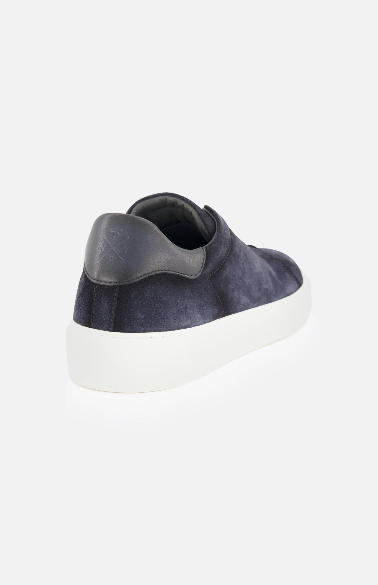 Presenting the James Slip On Sneaker by TO BOOT—a stylish navy blue suede slip-on with a white sole, showcased from the back left angle. The heel is adorned with a dark leather patch featuring embossed lettering, highlighting its expert Italian craftsmanship. This refined sneaker stands out against a plain white background.