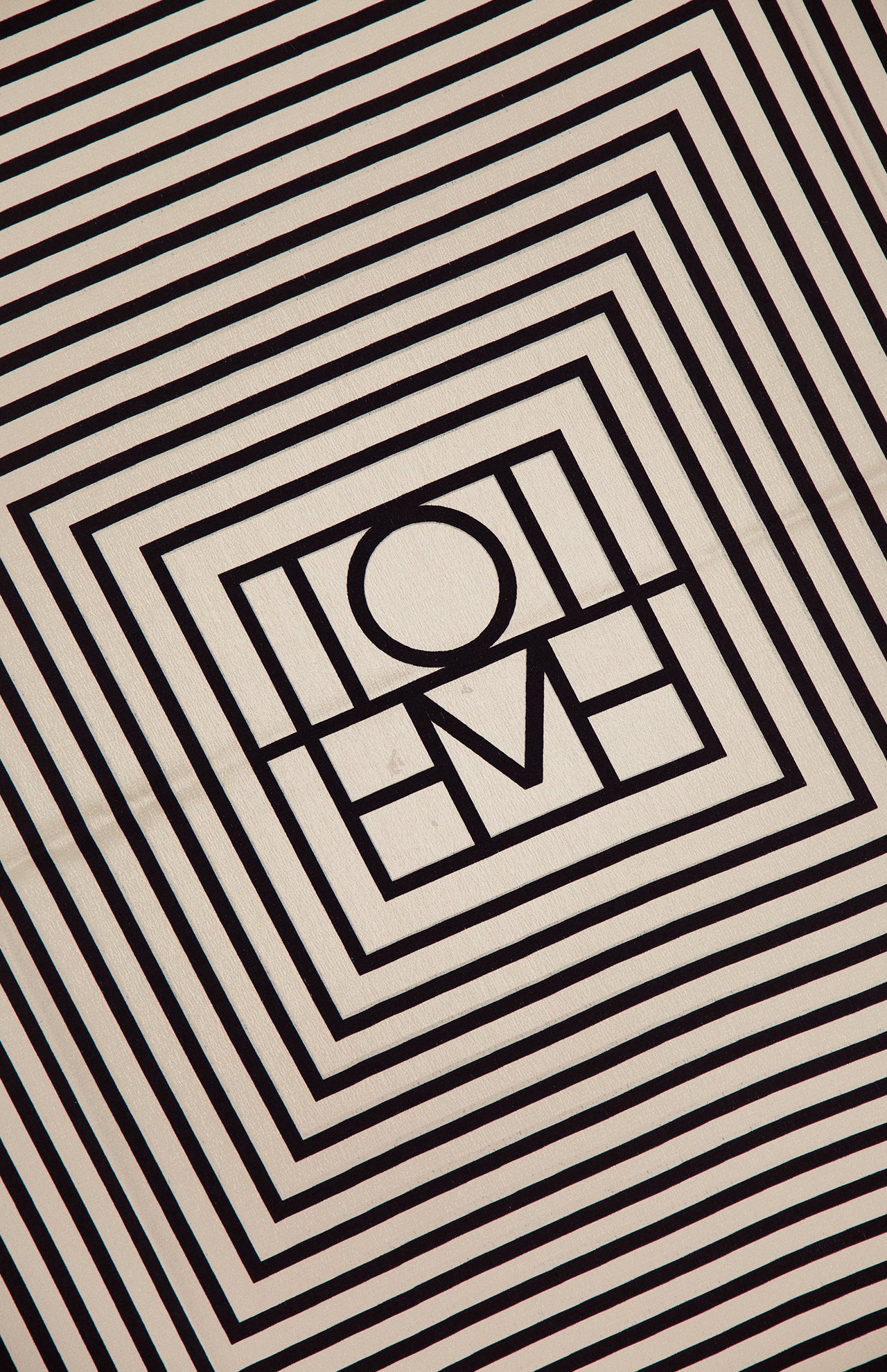 The TOTEME Centered Monogram Silk Scarf features black geometric lines forming concentric squares and a central HOME motif on beige silk, creating a bold optical illusion for women’s fashion.