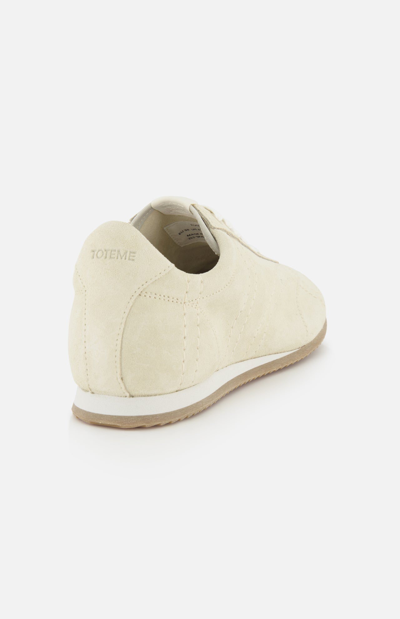 The Flex Suede Low Profile Sneakers by TOTEME feature beige minimalist suede uppers and light brown soles, viewed from the back/side. TOTEME is embossed on the heel. Made in Italy, shown against a white background.